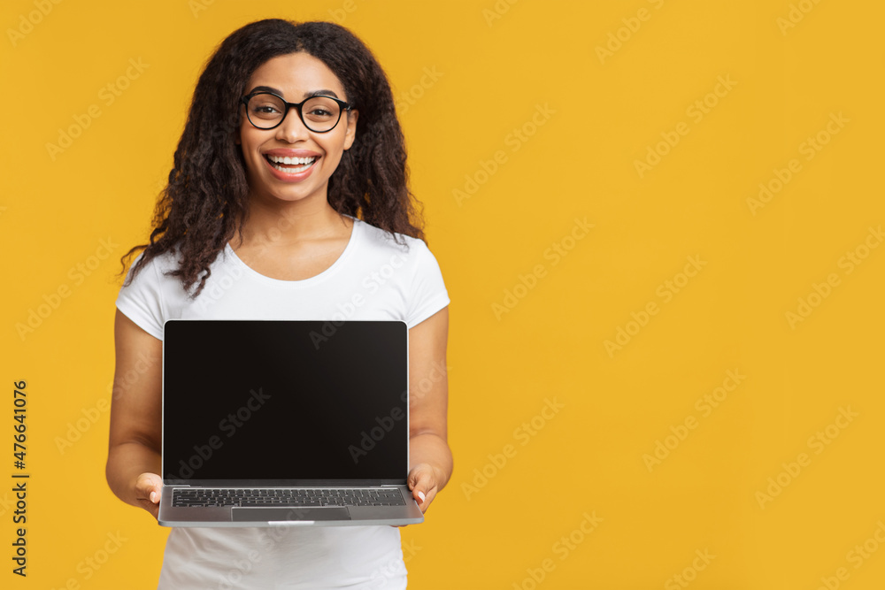 Excited black student demonstrating laptop with blank screen, standing over yellow studio background, empty space Excited black student demonstrating laptop with blank screen, standing over yellow studio background, empty space