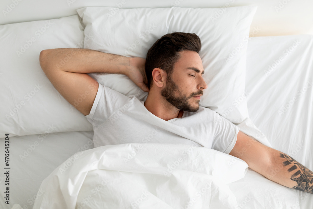 Tired millennial man sleeping, lying on white bed in bedroom, resting with eyes closed , top view, copy space Tired millennial man sleeping, lying on white bed in bedroom, resting with eyes closed , top view, copy space