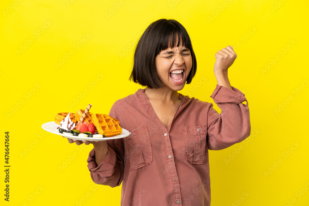 Pregnant woman holding waffles isolated on yellow background celebrating a victory