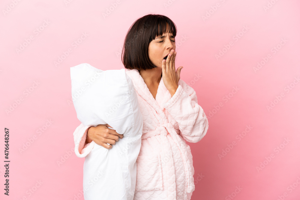 Young mixed race pregnant woman isolated on pink background in pajamas and holding a pillow and yawning Young mixed race pregnant woman isolated on pink background in pajamas and holding a pillow and yawning