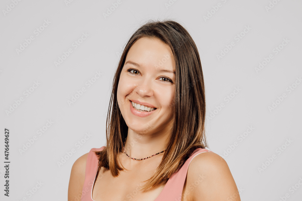 Portrait of brunette on isolated gray background. Young happy woman poses in studio, turning sideways and smiling, she is wearing beige top for yoga or fitness. Portrait of brunette on isolated gray background. Young happy woman poses in studio, turning sideways and smiling, she is wearing beige top for yoga or fitness.