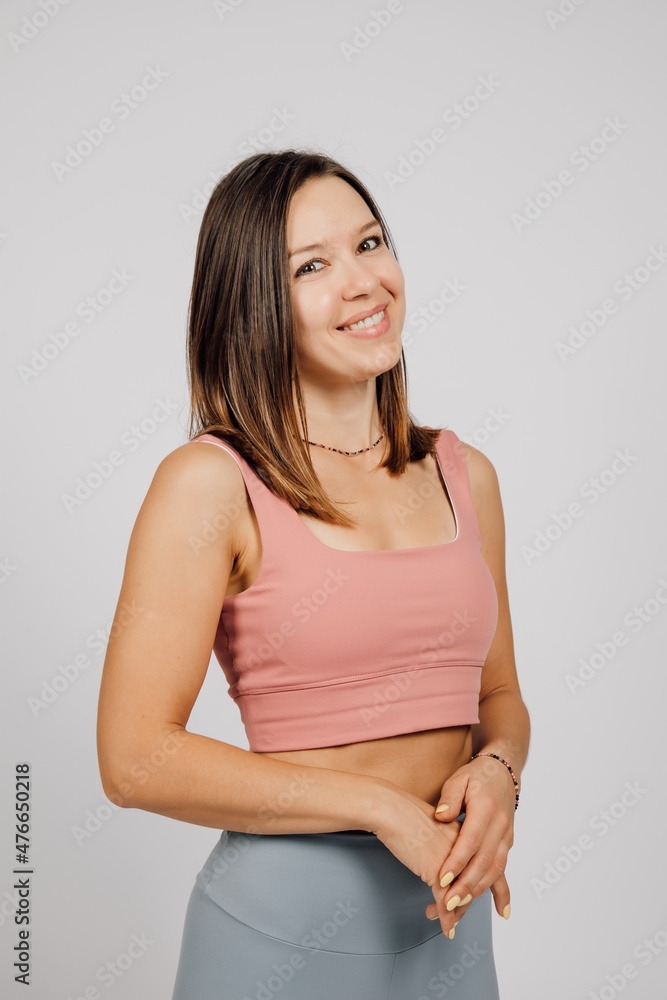 Beautiful woman posing in studio. Portrait of happy young brunette in beige top and gray yoga or fitness pants, she stands on gray isolated background and smiles, putting one hand on other. Beautiful woman posing in studio. Portrait of happy young brunette in beige top and gray yoga or fitness pants, she stands on gray isolated background and smiles, putting one hand on other.