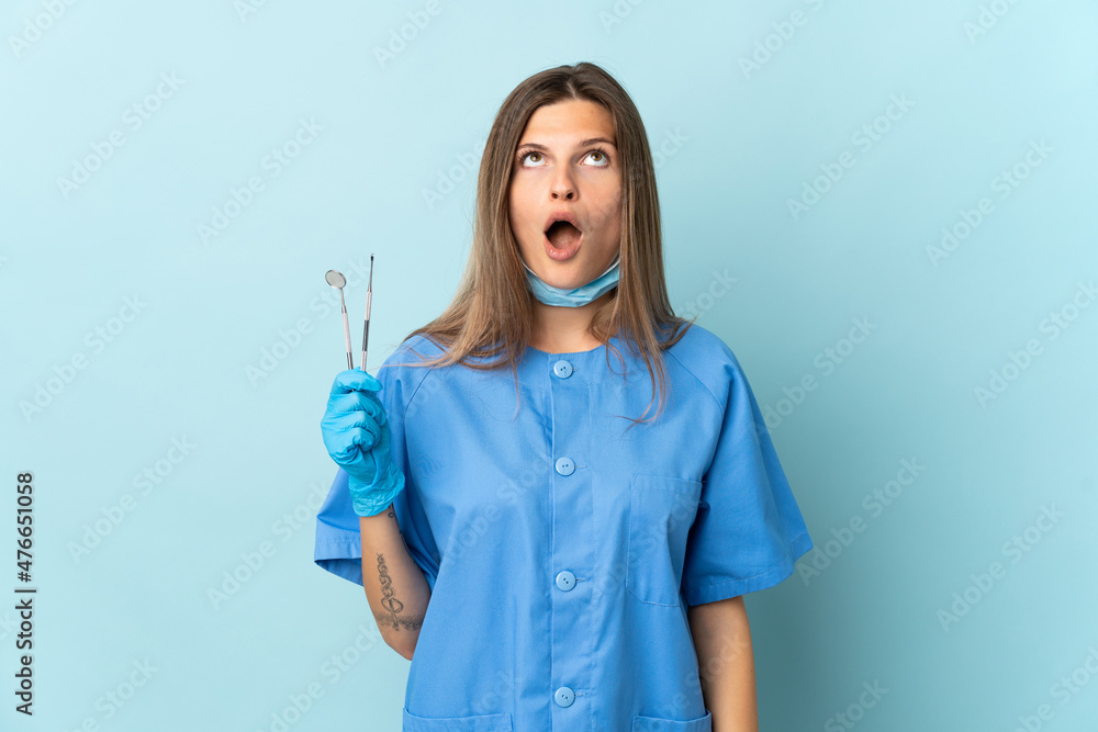 Slovak dentist holding tools isolated on blue background looking up and with surprised expression Slovak dentist holding tools isolated on blue background looking up and with surprised expression