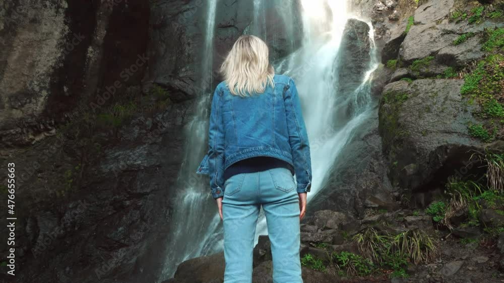 a blonde traveler in a denim jacket and jeans enjoys freedom, feeling happy, facing the waterfall and with her back to the camera. The wind ruffles her hair. Happy blonde girl. Makhuntseti, Georgia. a blonde traveler in a denim jacket and jeans enjoys freedom, feeling happy, facing the waterfall and with her back to the camera. The wind ruffles her hair. Happy blonde girl. Makhuntseti, Georgia.