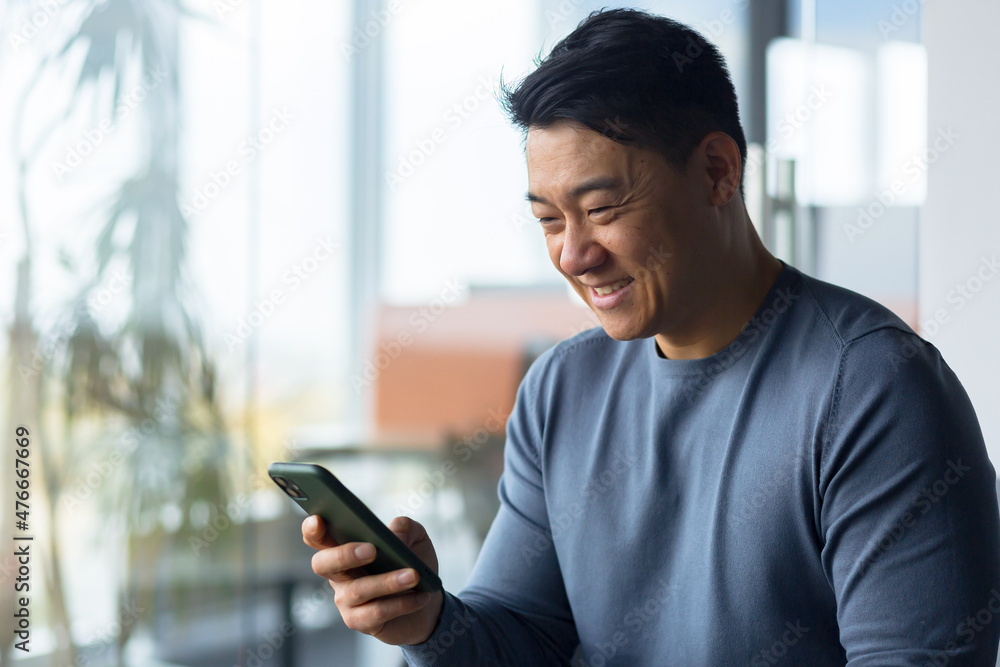 Close-up photo of male businessman using phone in modern office, asian looking at phone display and smiling, online call Close-up photo of male businessman using phone in modern office, asian looking at phone display and smiling, online call