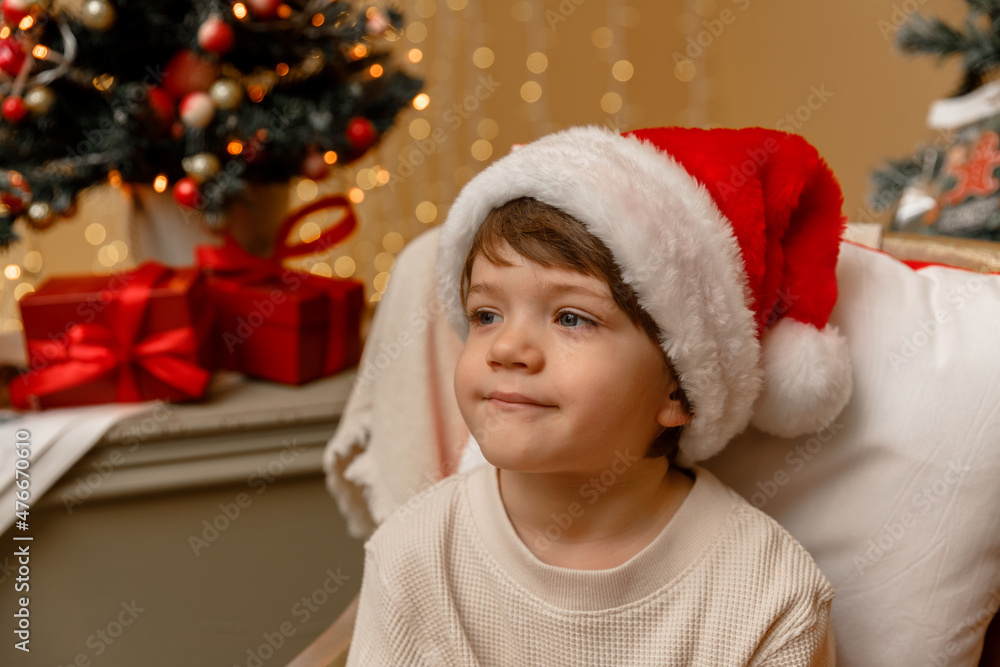 A child with Santa Claus hat sits in front of the Christmas tree A child with Santa Claus hat sits in front of the Christmas tree