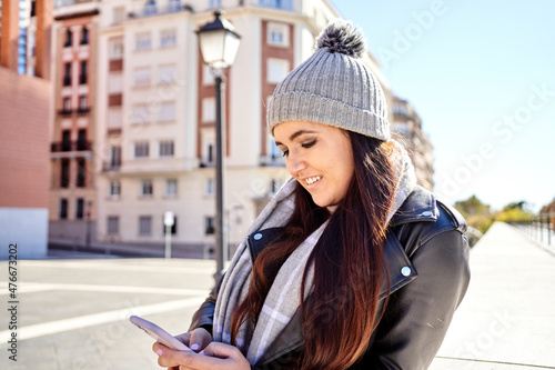 Side view of young smiling lady with long brown hair in trendy leather jacket and knitted hat messaging on mobile phone leaning on stone border, on city street © JoseIMartin