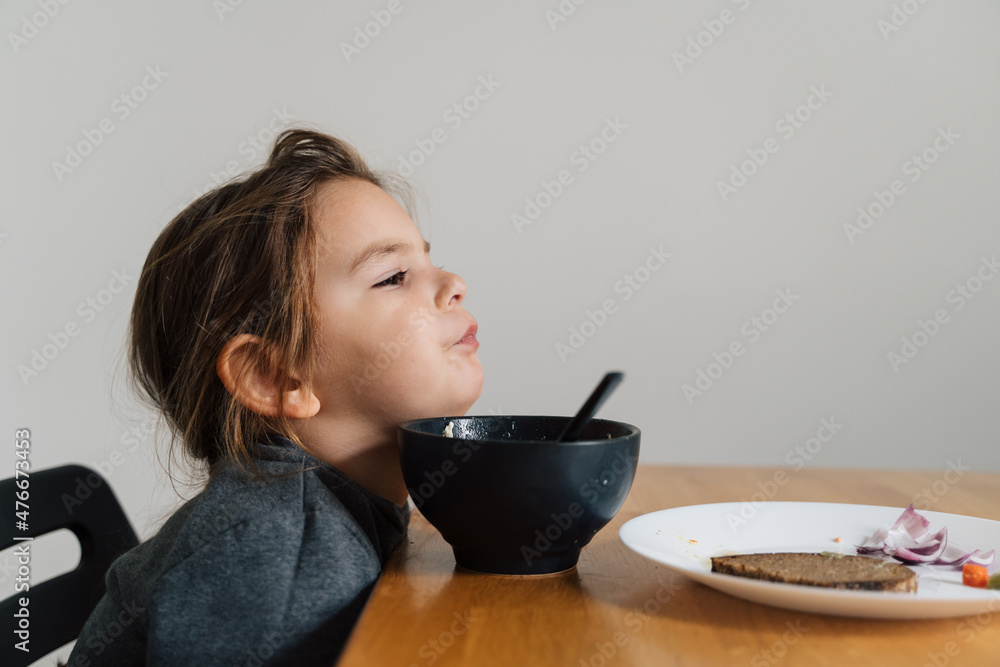 Unhappy child girl eats soup from black bowl with bread and onion. Lifestyle photo of kid in kitchen having a meal, Screaming kid. Picky eater Unhappy child girl eats soup from black bowl with bread and onion. Lifestyle photo of kid in kitchen having a meal, Screaming kid. Picky eater