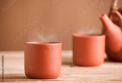Two brown ceramic cups with tea or coffee and hot steam, on rustic wooden table, partial view of teapot or coffee pot, brown background. Space for text.