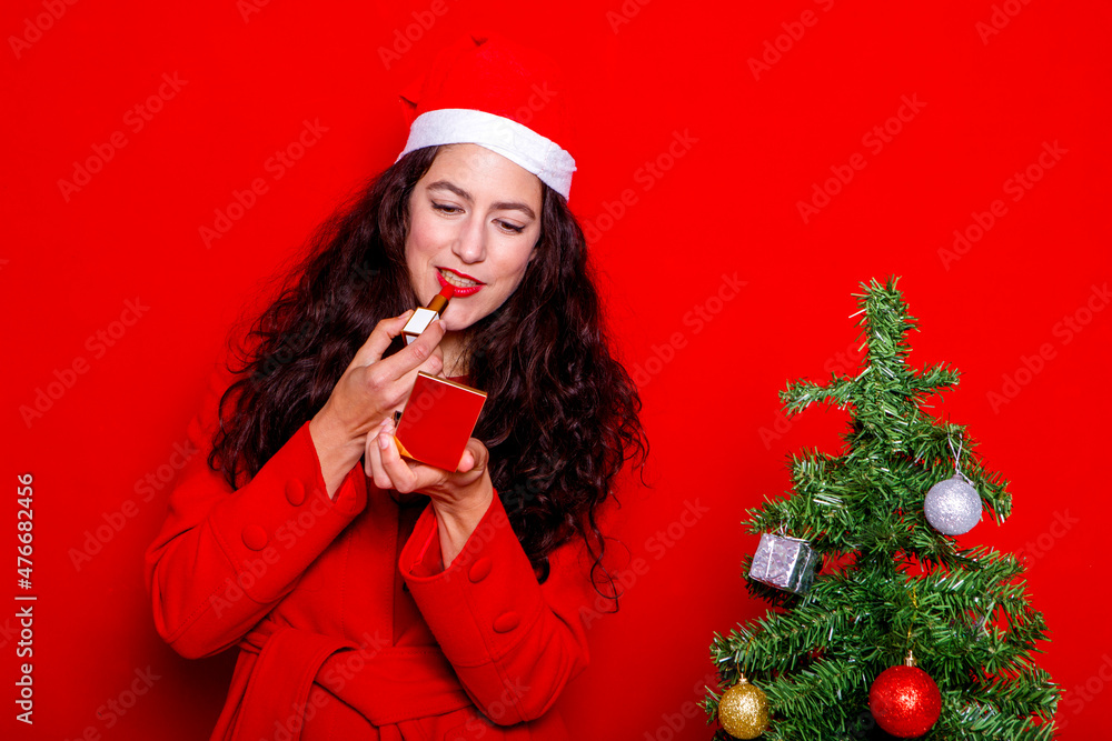 girl in red coat and christmas hat putting on makeup on red background girl in red coat and christmas hat putting on makeup on red background