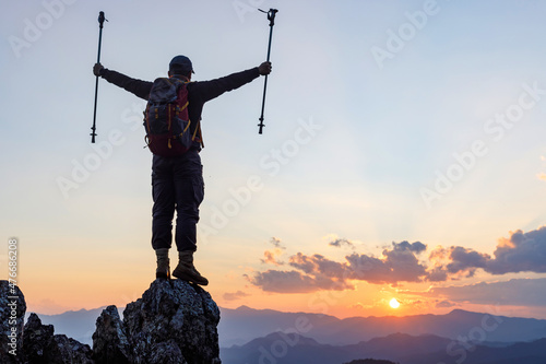 Silhouette Successful hiker outstretched arms with holding stick on mountain top cliff edge at sunset background. Success people and hiking adventure concept.