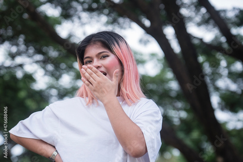 A happy young asian woman with pink highlights pretends to yawn showing boredom or an expression of being unimpressed. Outdoor scene.