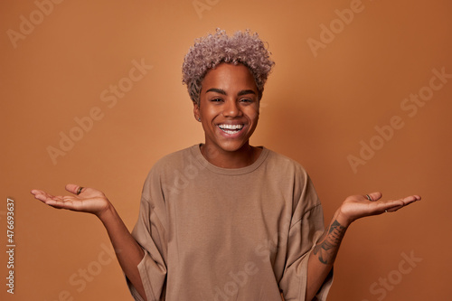 Surprised joyful young African American woman looks directly at you smiling broadly spreading hands, demonstrating incomprehension and indecision standing in casual clothes on brown background photo