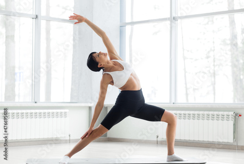 Woman n a white top and black bicycles practicing yoga, standing in anjaneyasana pose, girl doing Horse rider exercise on mat, working out at home or in yoga studio with white walls