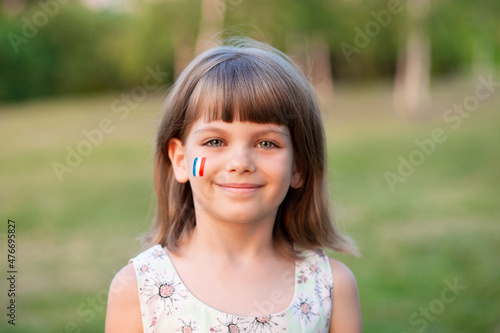 Outdoor portrait of little caucasian kid girl with cheeks painted in France flag colors and looking into camera and smile. Young painter. Modern Art.