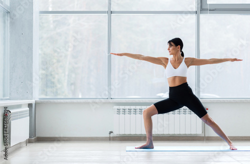 Young yogi woman practicing yoga, standing in Warrior two exercise, Virabhadrasana II pose, working out wearing sportswear bra and pants, full length, white loft studio background. Weight loss concept
