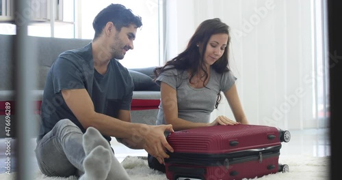 Content pregnant woman and man closing packed suitcase with baby clothes while sitting together on floor at home and preparing for maternity hospital