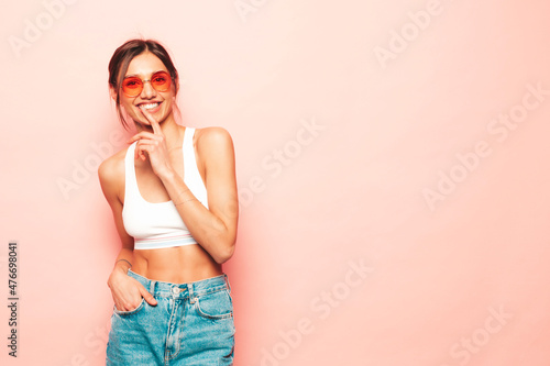 Beautiful smiling woman dressed in white jersey top shirt and jeans. Sexy carefree cheerful model having fun indoors.Adorable and positive female posing near pink wall in studio. In sunglasses