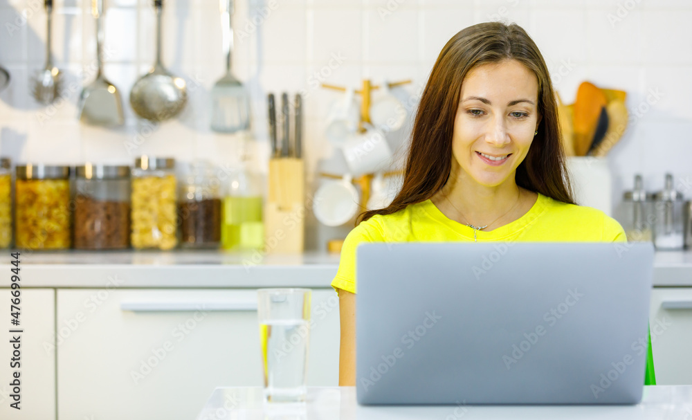 Front view of happy caucasian young pregnant female mother smiling and browsing data on netbook while sitting at table and working on remote project in kitchen at home Front view of happy caucasian young pregnant female mother smiling and browsing data on netbook while sitting at table and working on remote project in kitchen at home