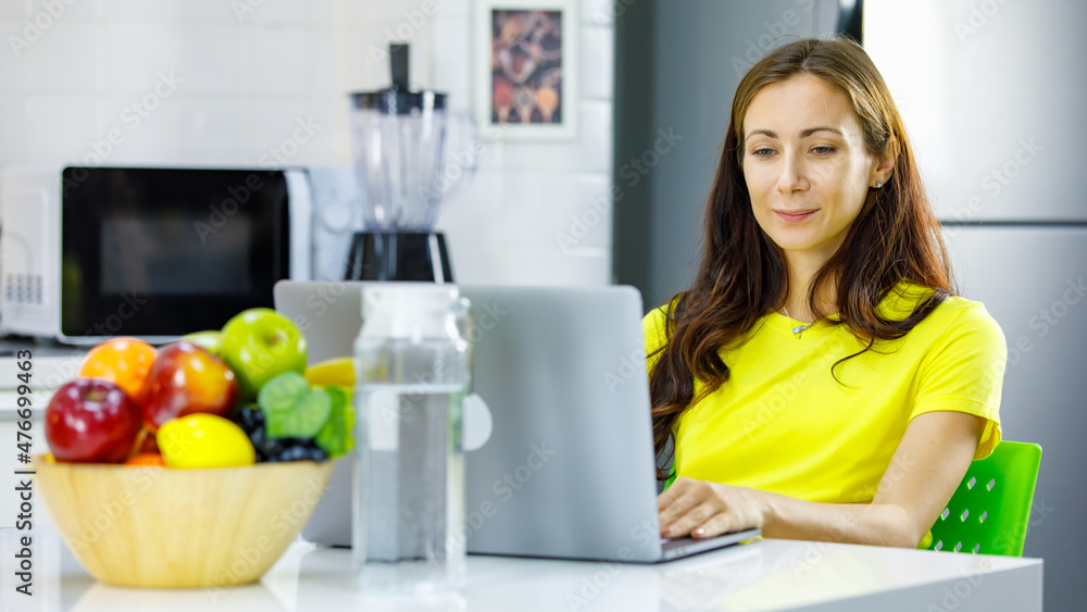 Front view of happy caucasian young pregnant female mother smiling and browsing data on netbook while sitting at table and working on remote project in kitchen at home