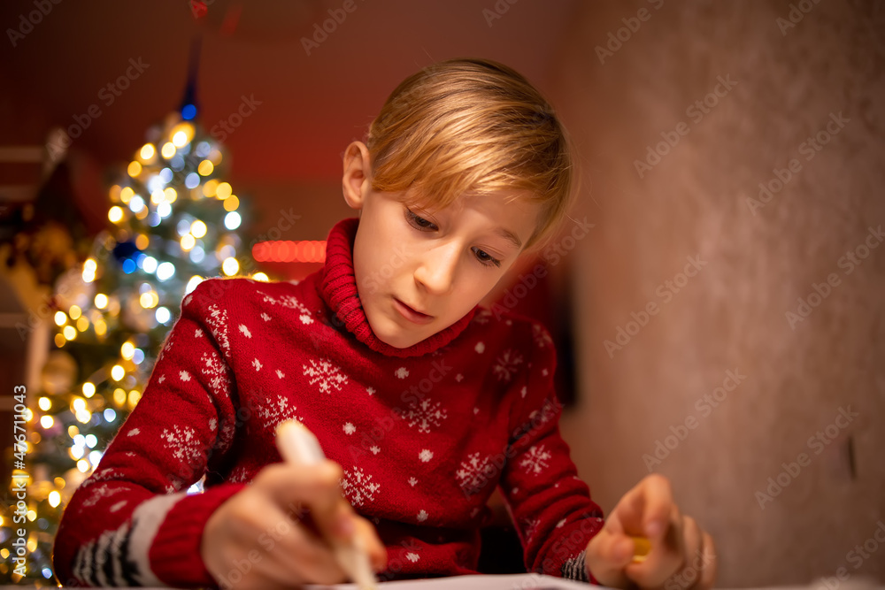 a boy in a red Christmas sweater on the background of a Christmas tree is engaged in the process of drawing in his room under a lamp a boy in a red Christmas sweater on the background of a Christmas tree is engaged in the process of drawing in his room under a lamp