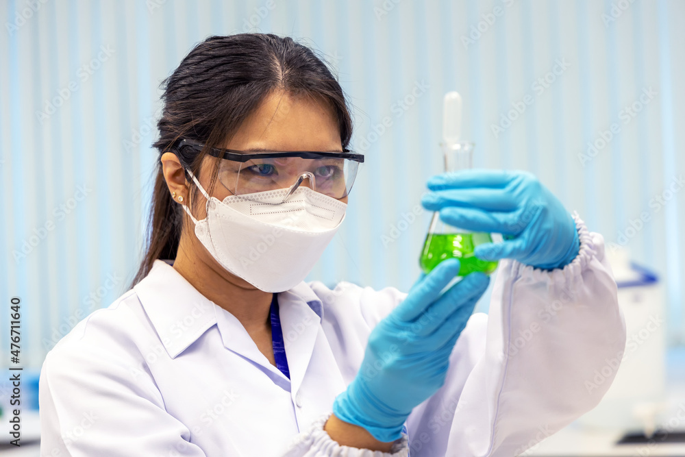 Woman scientist working in laboratory with Erlenmeyer flask with solutions in laboratory. Technician woman in medical coat uniform with protective goggles holding solvent liquid. Selective in face. Woman scientist working in laboratory with Erlenmeyer flask with solutions in laboratory. Technician woman in medical coat uniform with protective goggles holding solvent liquid. Selective in face.