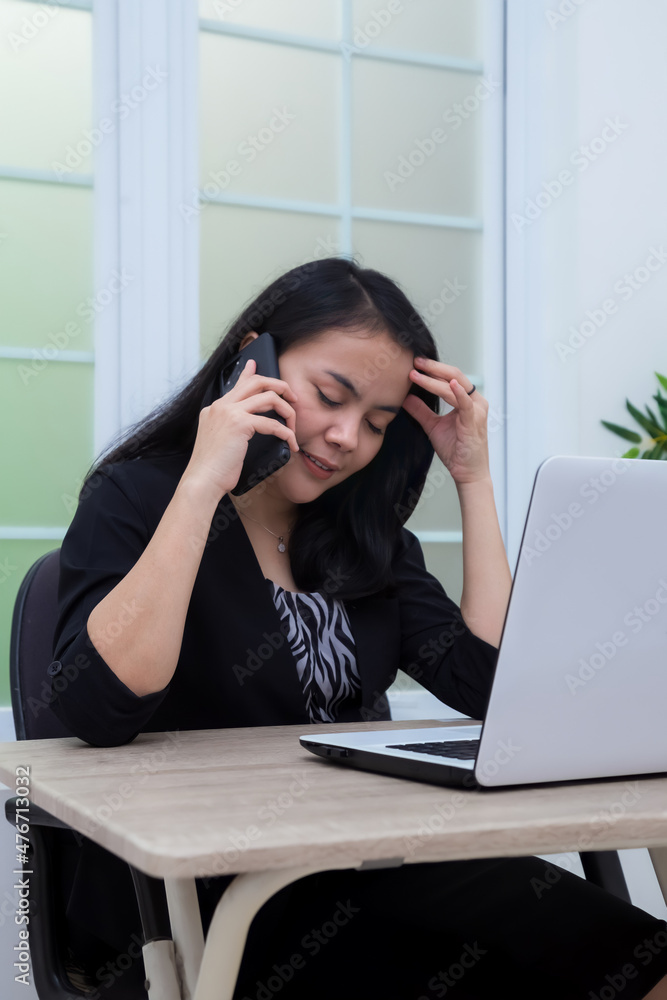 Business woman sitting on chair while making phone holding head in front of laptop Business woman sitting on chair while making phone holding head in front of laptop