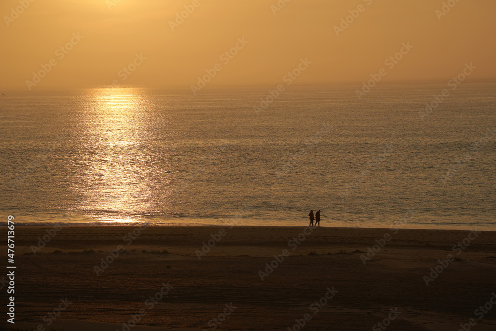 2 people walking on the North Sea beach in South Holland. Sunset over the orange shining sea. Togetherness.