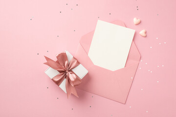 Top view photo of white giftbox with pink ribbon bow small hearts open pink envelope with paper card and shiny sequins on isolated pastel pink background with blank space