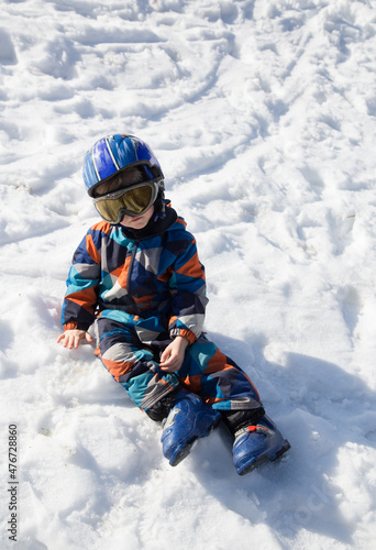 tired kid in a helmet, glasses, ski boots and winter overalls sits in the snow at a ski resort. Children's skiing lesson at an alpine school. Fell in the snow, pause to rest. Winter activities