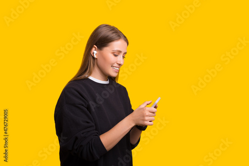 Young woman holding mobile cell phone listening to music with earpods isolated on yellow background studio