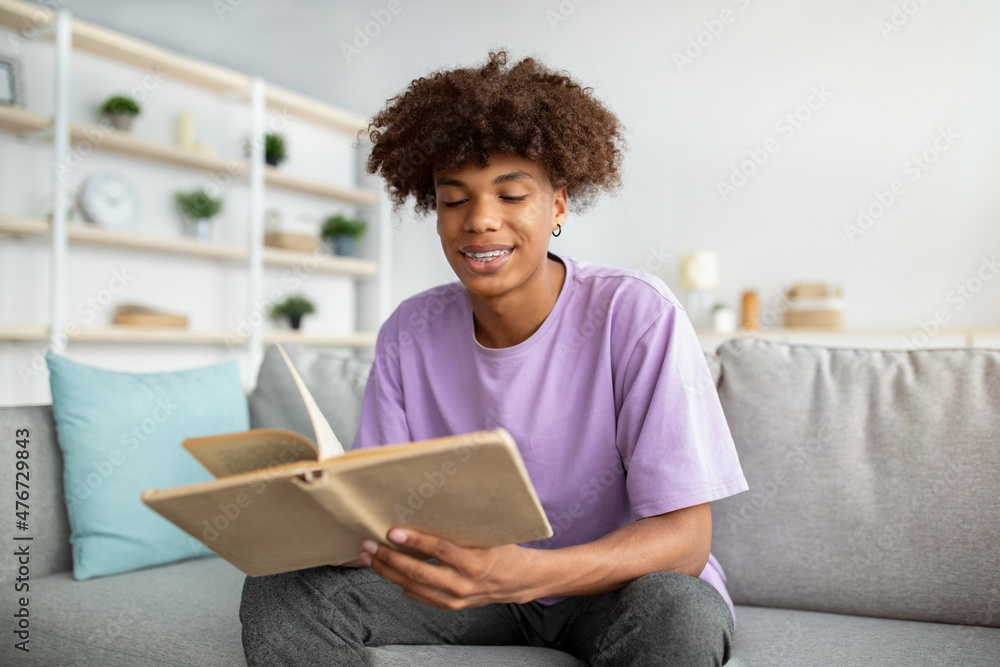 Positive teenage guy with open book sitting on comfy sofa, studying or doing homework indoors Positive teenage guy with open book sitting on comfy sofa, studying or doing homework indoors