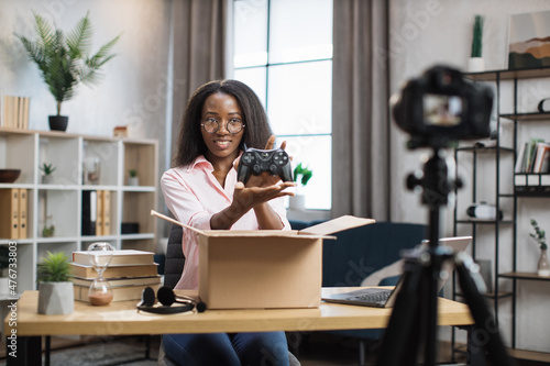 Positive young woman holding new wireless joystick while sitting at desk with opened cardboard box. Female customer examining purchase at home. Delivery concept.
