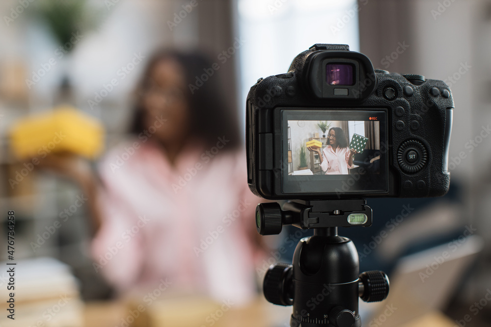 Close up of modern video camera with african american woman on screen. Female blogger unboxing holidays presents while sitting at desk. Blur background.
