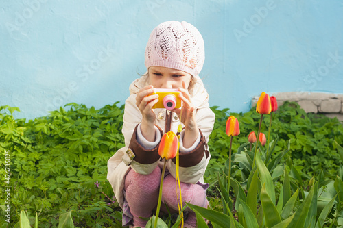 a girl in a white jacket sits with a small camera in front of a tulip