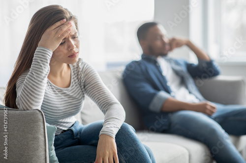 Upset Young Interracial Spouses Sitting On Couch, Ignoring Each Other After Quarrel
