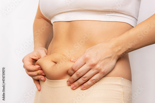 A cropped shot of a young woman standing sideways holding herself by the fat on her stomach isolated on a white background. Overeating, overweight, excess weight, hormonal belly