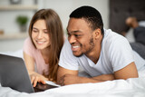 Affectionate multiracial couple spending time together at home, using laptop on bed