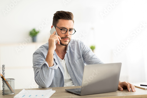 Handsome young man talking on smartphone, speaking with customer, working on laptop at home office workplace