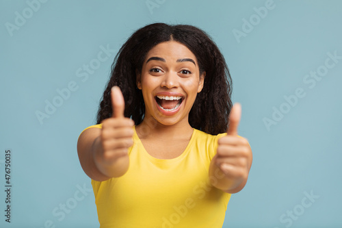 Good promo. Overjoyed black lady showing thumbs up with both hands and smiling to camera over blue background