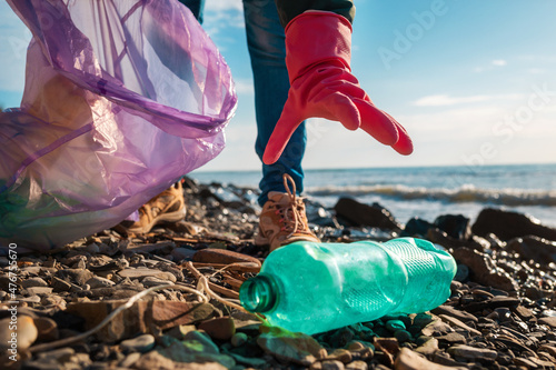 A volunteer in rubber gloves reaches for a dirty plastic bottle lying on the ocean shore. Hand close-up. The concept of environmental purification
