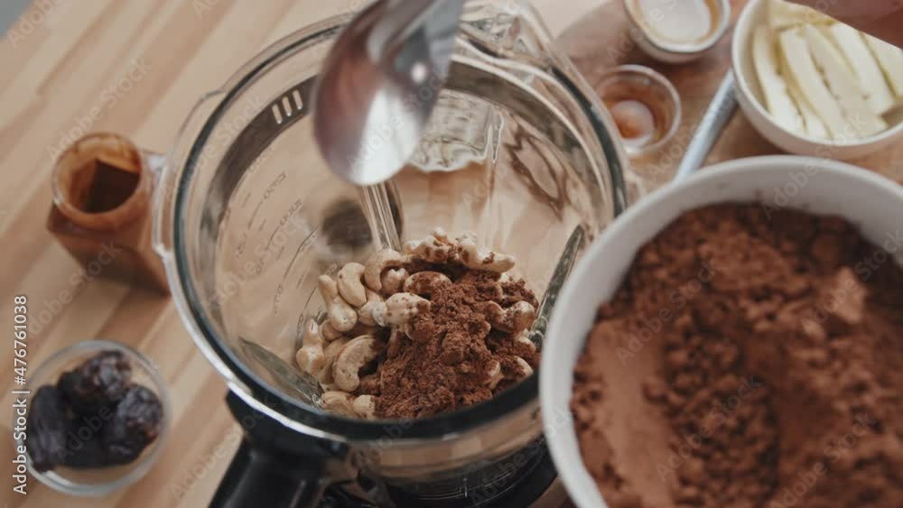 Handheld close up shot of unrecognizable woman adding cocoa to cashews in blender while making healthy smoothie in morning Handheld close up shot of unrecognizable woman adding cocoa to cashews in blender while making healthy smoothie in morning