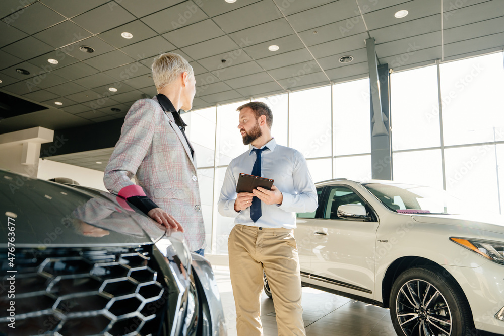 Salesman at the dealership showroom talking with customer and helping her to choosing a new car for herself in car showroom vehicle salon dealership store motor showroom. Salesman at the dealership showroom talking with customer and helping her to choosing a new car for herself in car showroom vehicle salon dealership store motor showroom.