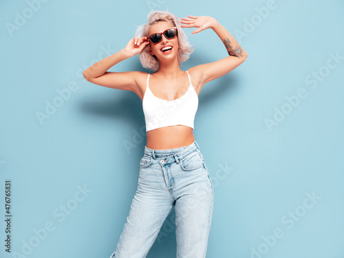 Young beautiful smiling female in trendy summer clothes. Sexy carefree woman posing near blue wall in studio. Positive blond model having fun and going crazy. In sunglasses