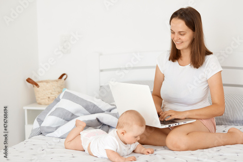 Positive smiling woman sitting on bed with her infant daughter and working online, holding laptop, looking at display, having video call or watching movie online.