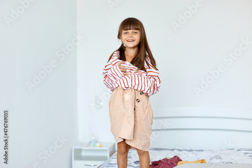 Portrait of small girl wearing large striped shirt and beige short pretending to be a grown up woman. Girl child posing in oversized clothes indoor while standing on bed.