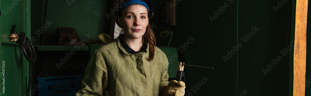Brunette welder looking at camera while holding welding torch in factory, banner Brunette welder looking at camera while holding welding torch in factory, banner