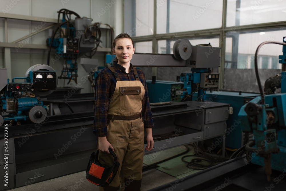 Young welder holding protective mask and looking at camera in factory