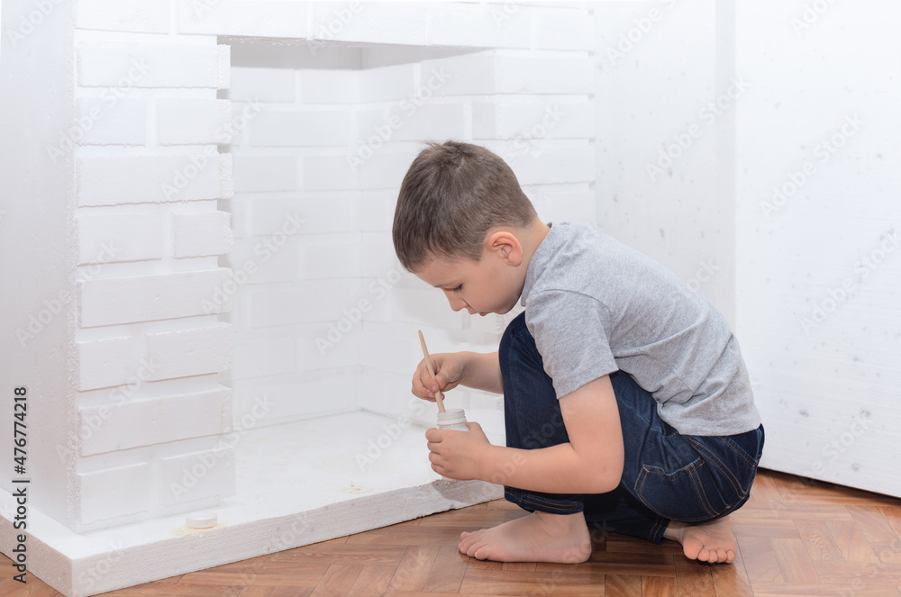 Child boy sits on the floor and paints a decorative fireplace with white paint gouache. Home improvement, DIY concept. Selective focus Child boy sits on the floor and paints a decorative fireplace with white paint gouache. Home improvement, DIY concept. Selective focus