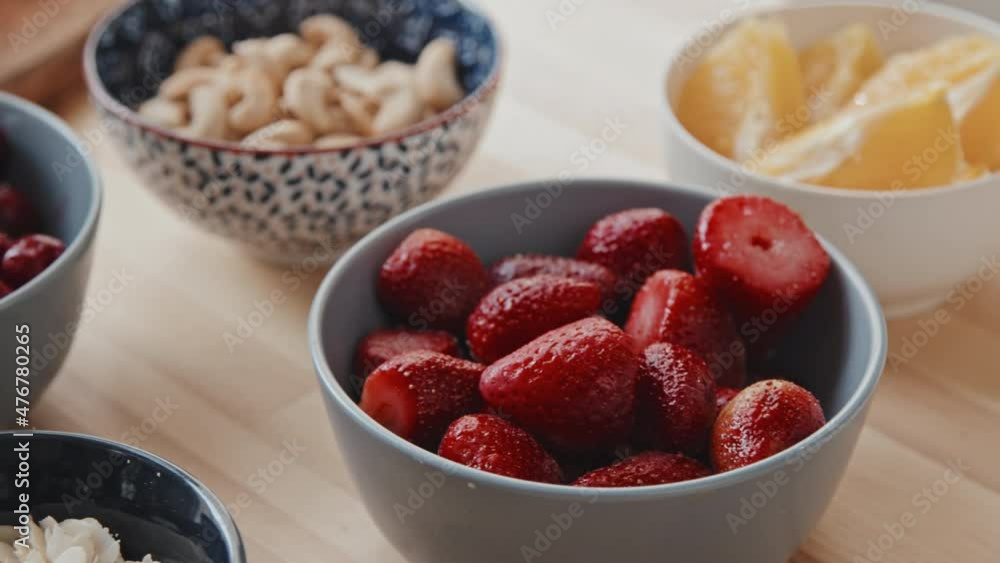 Handheld extreme close up tracking of variety of ingredients on kitchen table nuts, cherries, strawberries and orange slices in bowls and milk in jug Handheld extreme close up tracking of variety of ingredients on kitchen table nuts, cherries, strawberries and orange slices in bowls and milk in jug